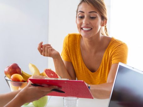Frau in orangenem Shirt lächelt und schaut auf ein Tablet. Ein Obstkorb steht neben ihr.