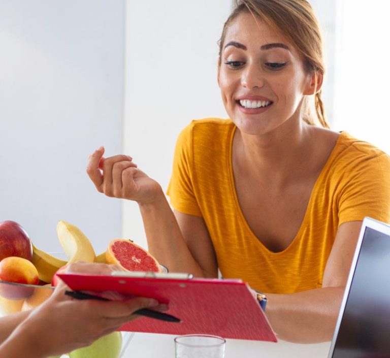 Junge Frau in orangenem Outfit lächelt und betrachtet ein Tablet an einem Tisch mit Obst.