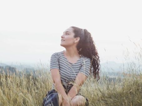 Frau mit lockigem Haar sitzt im Gras und schaut optimistisch in die Ferne.